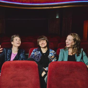 Drei lächelnde Personen sitzen in roten Kinosesseln: Links eine Frau mit kurzem Haar in glänzender Jacke, Mitte eine Person mit Pony in gemustertem Oberteil, rechts eine Frau mit langem Haar in grünem Blazer.