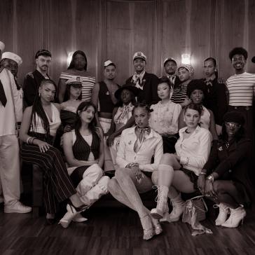 Gruppe von 18 Personen in maritimen und eleganten Outfits vor Holzvertäfelung: Matrosen, Offiziere und Personen in gestreifter Kleidung, stehend und hockend arrangiert.