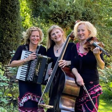 Drei lächelnde Frauen im Garten: Links spielt eine Akkordeon, Mitte hält einen Kontrabass, rechts spielt eine Geige, alle in schwarzen Outfits mit pinken Akzenten.