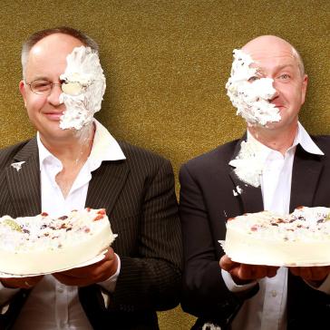 Zwei lächelnde Männer in Anzügen mit Krawatten halten jeweils eine Torte; Gesichter mit Sahne verschmiert, goldener Hintergrund.