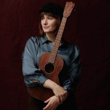 Lächelnde Person mit schwarzer Mütze und grauer Bluse hält eine braune Ukulele vor dunklem Hintergrund, Blick leicht zur Seite.
