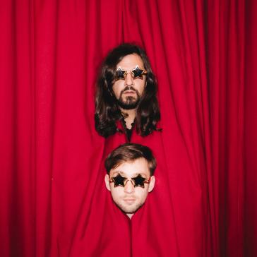 Zwei Personen stecken ihre Köpfe durch einen roten Bühnenvorhang: Beide tragen Sonnenbrillen in Sternenform, oben Person mit langen Haaren und Bart, unten Person mit kurzen Haaren.