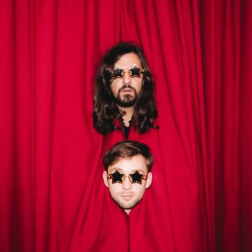 Zwei Personen stecken ihre Köpfe durch einen roten Bühnenvorhang: Beide tragen Sonnenbrillen in Sternenform, oben Person mit langen Haaren und Bart, unten Person mit kurzen Haaren.