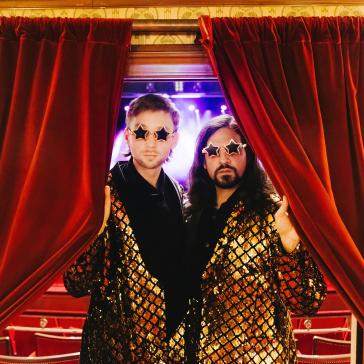 Zwei Personen mit glitzernden Jacken und Sternen-Sonnenbrillen posieren vor rotem Bühnenvorhang, eine hält den Vorhang auf, im Hintergrund blaue Bühnenbeleuchtung.