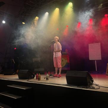 Person in rosa Shorts, weiß-gestreiftem Hemd und Basecap steht lächelnd mit Mikrofon auf der Bühne, bunte Scheinwerfer und Nebelmaschine, im Hintergrund Setlist auf Stehtafel.