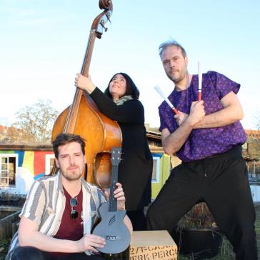 Drei Musiker:innen im Freien: Links sitzender Mann mit Ukulele, Mitte stehende Frau mit Kontrabass, rechts kniender Mann mit Stöcken vor einer Cajón-Kiste, bunte Häuser im Hintergrund.