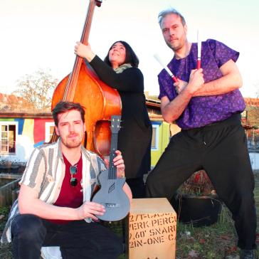 Drei Musiker:innen im Freien: Links sitzender Mann mit Ukulele, Mitte stehende Frau mit Kontrabass, rechts kniender Mann mit Stöcken vor einer Cajón-Kiste, bunte Häuser im Hintergrund.
