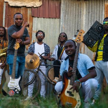 Sechs Musiker vor rostigem Wellblech: Links Saxophonist, Mitte Gitarrist, Bassist, Schlagzeuger mit Fahrradrahmen-Perkussion, rechts DJ mit Controller, alle lässig gekleidet, bunte Outfits.