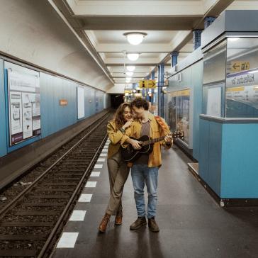 Zwei lächelnde Personen umarmen sich in einer U-Bahn-Station: Eine hält eine Gitarre, beide tragen lässige Kleidung, blaue Wandverkleidung und Gleise im Hintergrund.