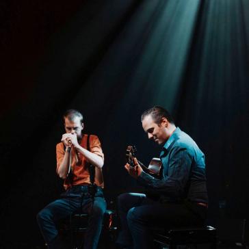 Zwei Musiker auf dunkler Bühne: Links sitzender Mann mit Mundharmonika und Mikrofon, rechts sitzender Mann spielt konzentriert Gitarre, dramatische Lichtstrahlen von oben.