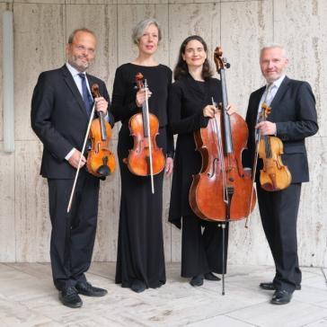 Vier Musiker:innen in schwarzer Kleidung vor hellem Steinmauer-Hintergrund: Zwei mit Violinen, eine mit Viola, einer mit Cello, alle Instrumente präsentierend.