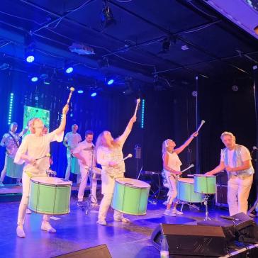 Sieben Musiker:innen in weißen Outfits spielen auf beleuchteter Bühne Trommeln und Perkussion, dynamische Posen, blaue Bühnenbeleuchtung und LED-Hintergrund.