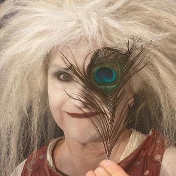Person mit wilden, weißen Haaren und theatralischem Make-up hält einen Pfauenbusch vor ein Auge, trägt gemustertes, rotes Oberteil und weiße Ärmel.