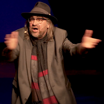 Mann mit Hut, Brille und rot-schwarz gestreiftem Schal steht mit ausgebreiteten Armen auf der Bühne, überraschter oder begeisterter Gesichtsausdruck, dunkler Hintergrund.