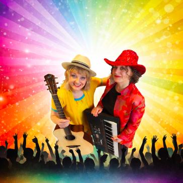 Zwei lächelnde Musikerinnen auf der Bühne: Links Frau mit Strohhut und Gitarre in gelbem Oberteil, rechts Frau mit rotem Hut und Keyboards, jubelndes Publikum und Regenbogen-Lichtstrahlen im Hintergrund.