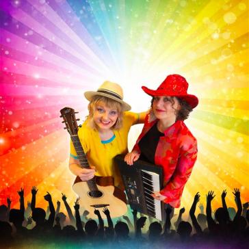 Zwei lächelnde Musikerinnen auf der Bühne: Links Frau mit Strohhut und Gitarre in gelbem Oberteil, rechts Frau mit rotem Hut und Keyboards, jubelndes Publikum und Regenbogen-Lichtstrahlen im Hintergrund.