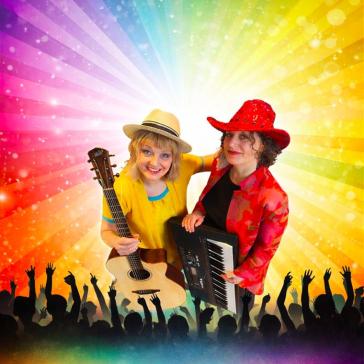 Zwei lächelnde Musikerinnen auf der Bühne: Links Frau mit Strohhut und Gitarre in gelbem Oberteil, rechts Frau mit rotem Hut und Keyboards, jubelndes Publikum und Regenbogen-Lichtstrahlen im Hintergrund.