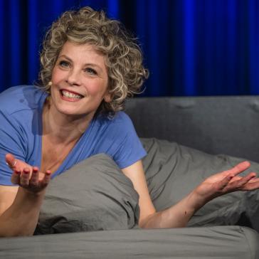 Lächelnde Person mit lockigen Haaren liegt entspannt in grauem Pyjama auf einem Bett, blaue Bettdecke, Arme ausgestreckt, als würde sie eine Geschichte erzählen, blauer Vorhang im Hintergrund.