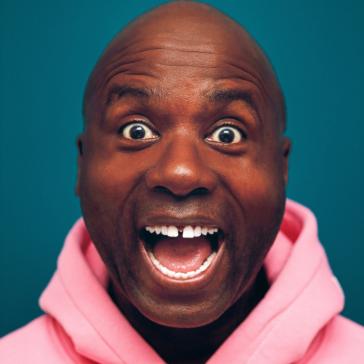 Lachelnde Person mit Glatze, weit geöffneten Augen und Mund, trägt einen rosa Kapuzenpulli, dunkler Hintergrund.