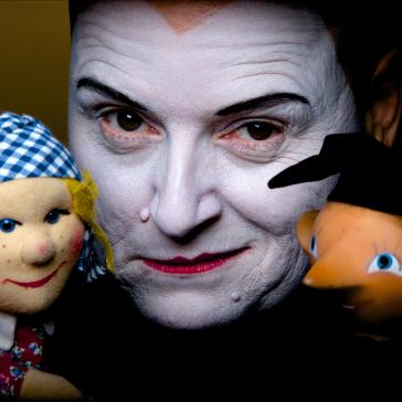 Person mit weißem Clown-Makeup, schwarzem Augenstreif, hält zwei Puppen: Links eine mit blau-kariertem Kopftuch, rechts eine mit orangefarbenem Haar und großen Augen.