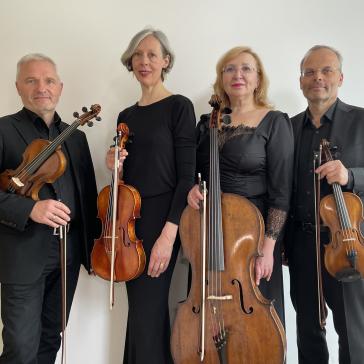 Vier lächelnde Musiker:innen in schwarzer Kleidung: Zwei mit Violinen, eine mit Cello, eine mit Bratsche, alle halten ihre Instrumente, helle Wand im Hintergrund.