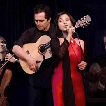 Vier Musiker auf der Bühne: Links Geiger, Mitte Gitarrist, Sängerin in rotem Kleid mit Mikrofon, rechts Schlagzeuger, alle konzentriert und in schwarzer Kleidung, dunkler Hintergrund.