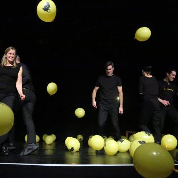 Fünf Personen tanzen auf dunkler Bühne zwischen vielen gelben Luftballons mit Hahnen-Aufdruck, alle in schwarzer Kleidung, dynamische Posen, fröhliche und lebendige Atmosphäre.
