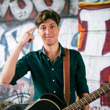 Lächelnder Mann mit dunklem Hemd und Gitarrenriemen hält eine schwarze Gitarre, posiert vor bunter Graffiti-Wand, Hand an der Schläfe, entspannte und kreative Atmosphäre.