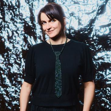 Lächelnde Person in schwarzem Pullover und Rock, mit glitzernder Kette als Krawattenersatz, steht vor funkelndem, beleuchtetem Hintergrund, Hände in den Taschen, kurze Haare.