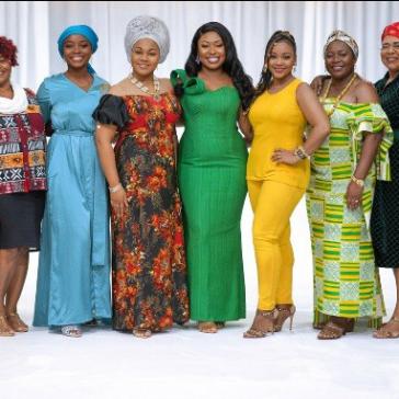 Sieben lächelnde Personen auf einem Laufsteg, alle in farbenfrohen, traditionellen afrikanischen Gewändern, verschiedene Muster und Farben, selbstbewusste Posen, Publikum im Hintergrund.