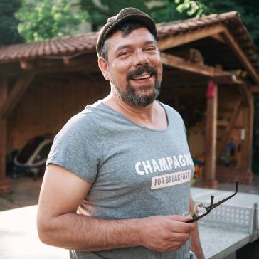 Mann mit Schiebermütze und T-Shirt hält seine Brille in der Hand und lacht und steht dabei vor einer Tischtennisplatte und einem Carport