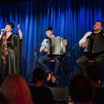 Vier Musiker auf der Bühne mit blauem Vorhang: Frau in schwarzem Kleid singt ins Mikrofon, links Saxophonist, rechts zwei Akkordeonspieler auf Hockern, Publikum im Vordergrund.