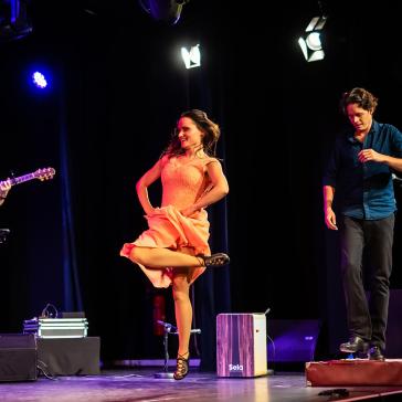 Drei Musiker auf der Bühne: Links Mann mit Gitarre, Mitte Frau in orangefarbenem Kleid tanzt, rechts Mann in blauem Hemd am Mikrofon, im Hintergrund blaue Vorhänge und Scheinwerfer.