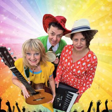 Drei Musiker auf der Bühne: Links Frau mit Gitarre in gelbem Kleid, Mitte Mann mit Tamburin in grünem Anzug und Sombrero, rechts Frau mit Keyboard in rotem Musterkleid, jubelndes Publikum im Hintergrund.