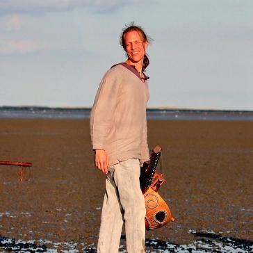 Drei Musiker am Strand: Links Mann mit traditionellem Instrument in orangefarbenem Gewand, Mitte Mann mit Gitarre in grauer Kleidung, rechts Frau mit Geige in weißem Kleid, Meer im Hintergrund.