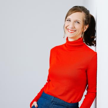 Frau mit Pferdeschwanz, rotem Rollkragenpullover und blauer Jeans lehnt lächelnd an weißer Wand, Hände in den Taschen, natürliches Licht.