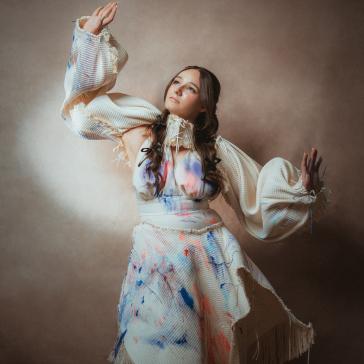 Frau in weißem, zerrissenem Kleid mit blauen Farbspritzern, weit ausgebreiteten Armen, dramatischer Spotlight-Effekt, dunkler Hintergrund.