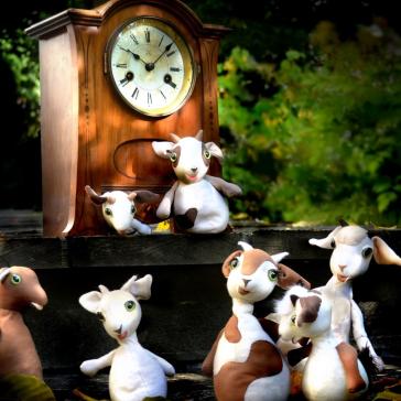 Sechs Stoff-Ziegenfiguren vor altem Holzwecker mit weißem Ziffernblatt, eine Ziege sitzt auf der Uhr, grüner Gartenhintergrund mit Blättern.