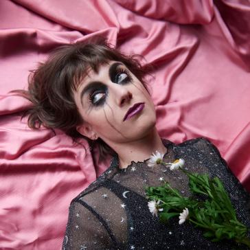 Frau mit dramatischem Make-up, schwarzem Sternenkleid und Gänseblumen im Haar liegt auf pinkem Satin, dunkles Haar, blauer Lidschatten, ernster Blick.