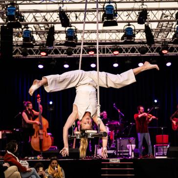 Artistin in weißem Kostüm hängt kopfüber an Trapez, Publikum im Vordergrund, im Hintergrund Band mit Kontrabass, Geige und Gitarre auf beleuchteter Bühne.