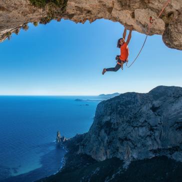 Person in orangefarbenem Shirt klettert an Felswand über türkisfarbenem Meer, Seil in der Hand, spektakuläre Aussicht auf Klippen und Horizont.