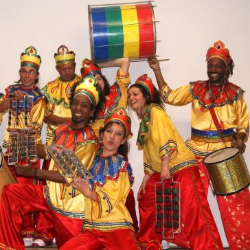 Acht Musiker in gold-rot-blauen Kostümen mit buntem Kopfschmuck, posierend mit Trommeln, Rasseln und einer grün-gelb-roten Trommel.