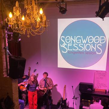 Live-Auftritt bei ‚Songwood Sessions‘: Drei Musiker mit Gitarre, Bass und Mikrofon auf kleiner Bühne, lila Beleuchtung, goldener Kronleuchter im Hintergrund.