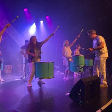 Sechs Musiker in weißen Outfits spielen grüne Trommeln auf einer pink beleuchteten Bühne, einige halten Schlägel in der Hand.