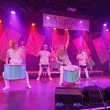 Sieben Musiker in weißen Outfits spielen Trommeln und Becken auf einer pink beleuchteten Bühne mit transparenten Vorhängen.