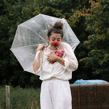 Clown mit roter Nase hält durchsichtigen Regenschirm und pinken Ballon, trägt weißes Outfit, steht im Regen vor grünem Waldhintergrund.
