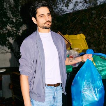 Mann mit grauer Jacke, weißem T-Shirt und Jeans hält blaue Mülltüte, im Hintergrund Müllcontainer und grüne Büsche.