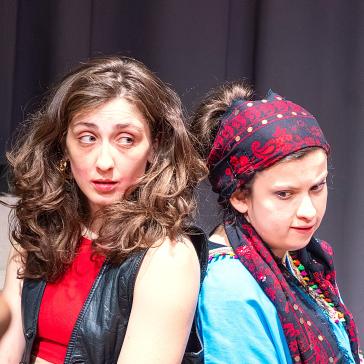 Zwei Frauen vor dunklem Hintergrund: Links mit lockigem Haar, roter Bluse und schwarzer Weste, rechts mit rot-schwarzem Tuch und blauem Oberteil.