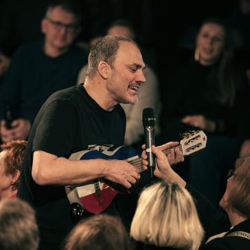 Mann in schwarzem Shirt spielt blaue Gitarre und singt ins Mikrofon, umgeben von aufmerksamen Zuhörern in dunklem Raum.
