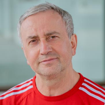 Mann mit graumeliertem Haar und Bart, trägt rotes Sportshirt mit weißen Streifen, vor hellem, unscharfem Hintergrund.